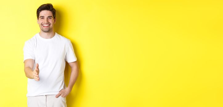 Handsome and confident man extending hand for handshake, greeting you, saying hello, standing in white t-shirt over yellow background.