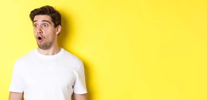 Close-up of impressed man looking left, gasping amazed, standing in white t-shirt against yellow background.