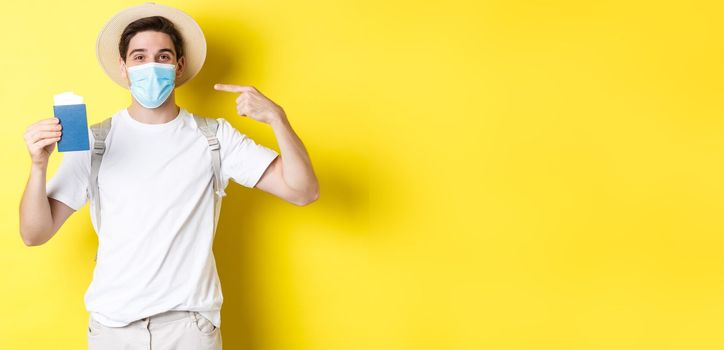 Concept of covid-19, tourism and pandemic. Man tourist showing passport, travelling in medical mask for protection from coronavirus, yellow background.