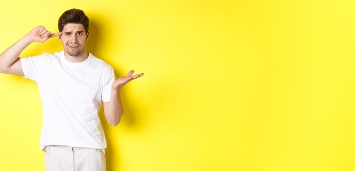 Confused and angry man pointing at head, scolding person for acting stupid, show crazy sign, standing over yellow background.