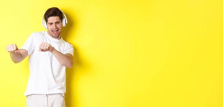 Cool guy listening music in headphones and dancing, standing in white clothes against yellow studio background.