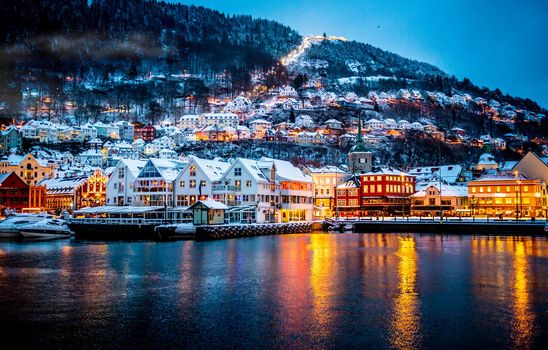Winter Bergen city with famous Bryggen merchandise wooden houses and lights in snow season. Panorama of historical harbor buildings at Christmas time with magical reflection in sea