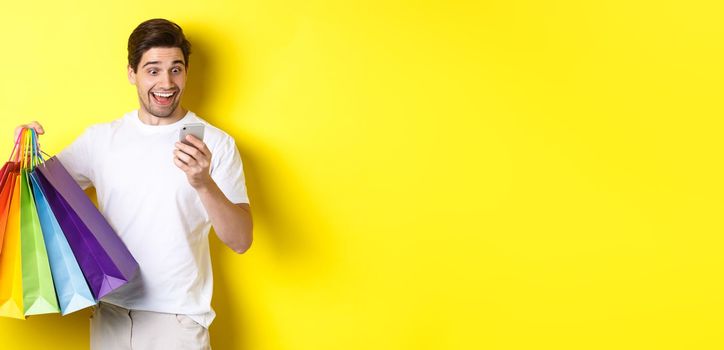 Excited man holding shopping bags and looking happy at mobile phone screen, standing over yellow background.