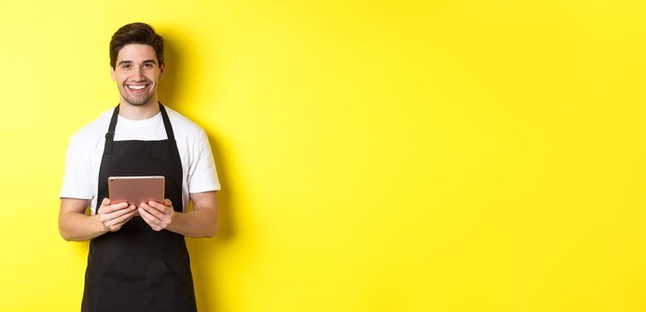 Waiter in black apron taking orders, holding digital tablet and smiling friendly, standing over yellow background.