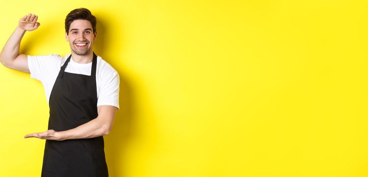 Smiling handsome barista showing something long or large, standing over yellow background.