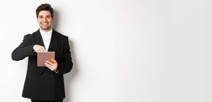 Portrait of handsome, stylish male entrepreneur in black suit pointing at digital tablet, showing something online, standing against white background.