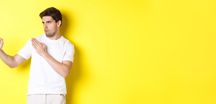 Man showing kung-fu skills, martial arts ninja movement, standing in white t-shirt ready to fight, standing over yellow background.