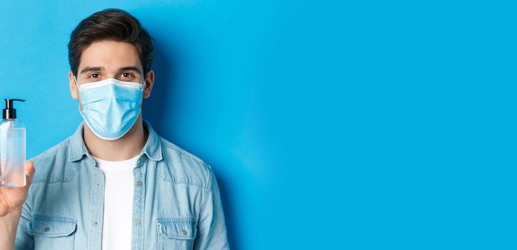 Concept of coronavirus, pandemic and preventive measures. Close-up of young man in medical mask advice to use hand sanitizer, showing antiseptic bottle, standing over blue background.