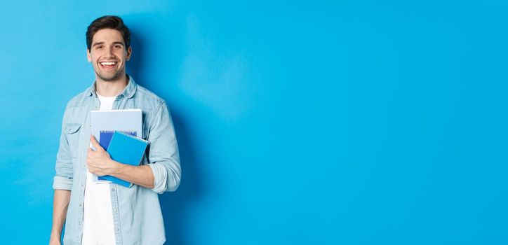 Smiling man studying, holding notebooks and looking happy, standing over blue background.