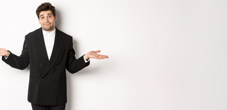Image of handsome confused man in black suit, shrugging and looking clueless, dont know anything, standing over white background.