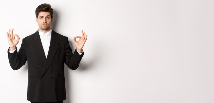 Concept of new year party, celebration and lifestyle. Portrait of confident good-looking man in black suit, showing okay sign and approve something, standing over white background.