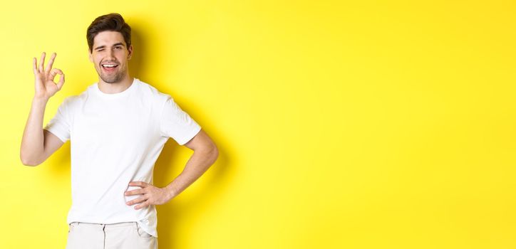 Confident handsome man winking, showing okay sign in approval, like something good, standing over yellow background.