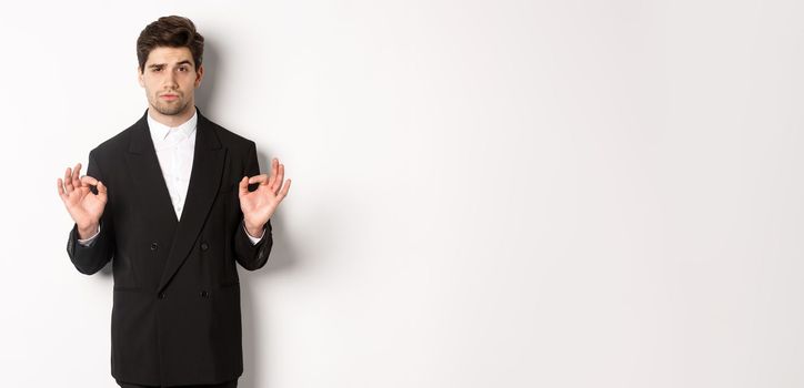 Concept of new year party, celebration and lifestyle. Portrait of confident good-looking man in black suit, showing okay sign and approve something, standing over white background.