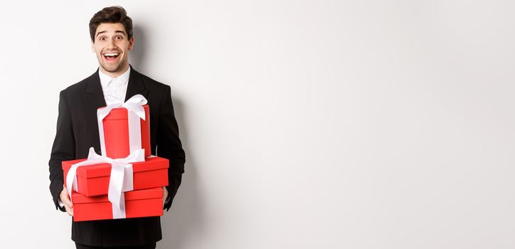 Concept of holidays, relationship and celebration. Handsome man in black suit bringing presents at new year party, holding gifts and smiling amused, standing against white background.