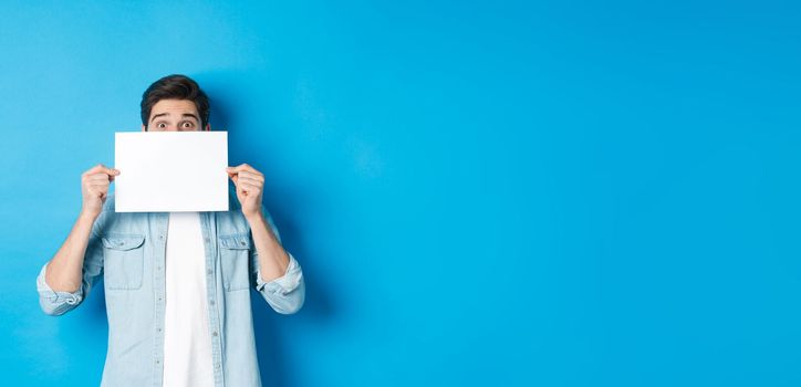 Excited man hiding face behind blank paper, place for your logo, standing over blue background.