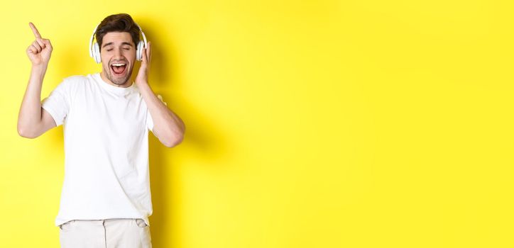 Happy man listening music in headphones, pointing finger at promo offer for black friday, standing over yellow background.