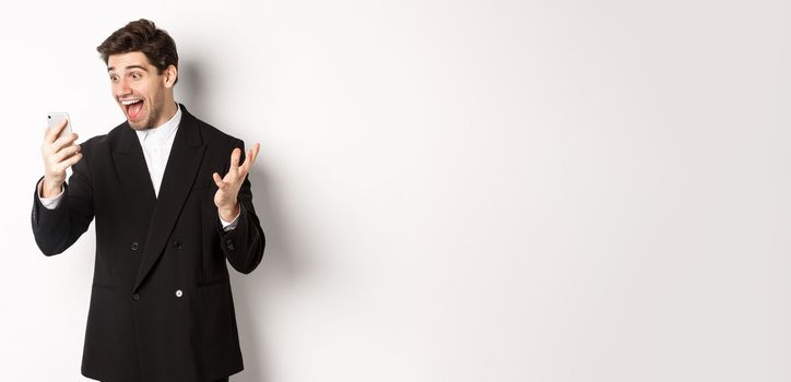 Portrait of happy smiling man in black suit, looking at smartphone screen with relieved and cheerful face, standing against white background.