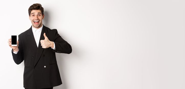 Image of handsome male entrepreneur in black suit, recommending app or online shop, showing thumbs-up and smartphone screen, standing over white background.