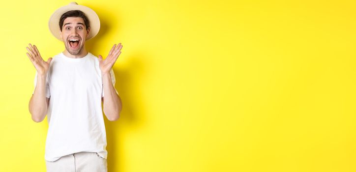 Concept of tourism and summer. Happy young man in straw hat looking amazed, reacting to surprise, standing over yellow background.