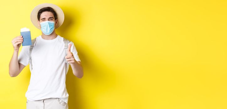 Concept of covid-19, tourism and pandemic. Happy guy tourist in medical mask showing passport with tickets for vacation, winking and pointing at camera, yellow background.