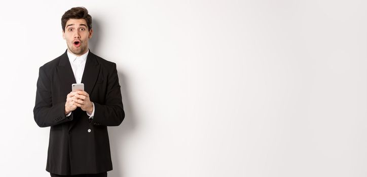 Amazed handsome guy in black suit reacting to cool promo offer, holding mobile phone, standing against white background.