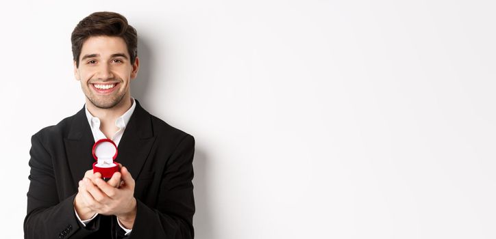 Image of handsome man looking romantic, open small box with engagement ring, making a proposal and smiling, standing against white background.