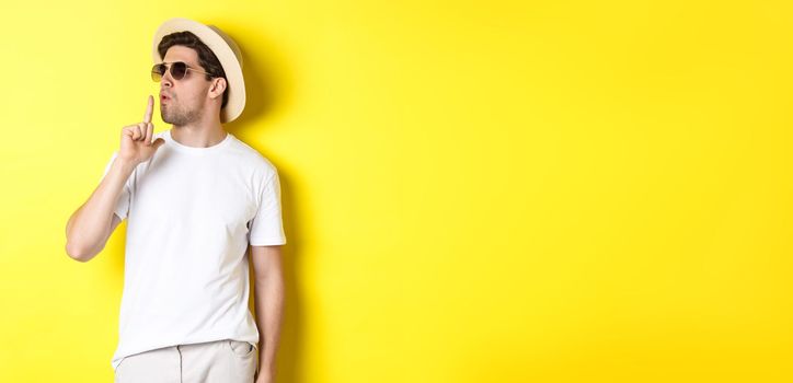 Cool young male tourist blowing at finger gun and looking confident, standing against yellow background. Vacation and lifestyle concept.