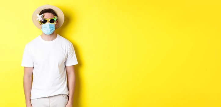 Concept of covid-19, vacation and social distancing. Man tourist wearing medical mask and summer hat with sunglasses, going on trip during pandemic, yellow background.