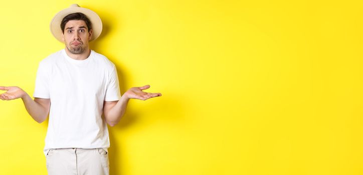 Concept of tourism and summer. Confused male tourist shrugging, looking indecisive, standing against yellow background.