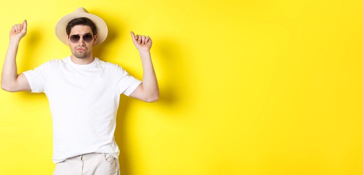 Tourism, travelling and holidays concept. Man tourist enjoying vacation, dancing in straw hat and sunglasses, posing against yellow background.