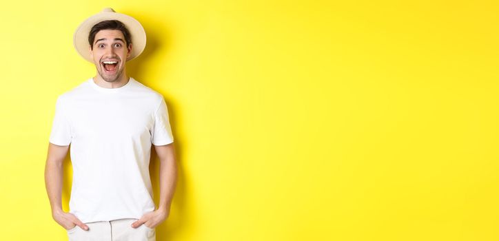 Surprised man tourist in straw hat looking happy, react amazed to travel agency advertisement, standing over yellow background.