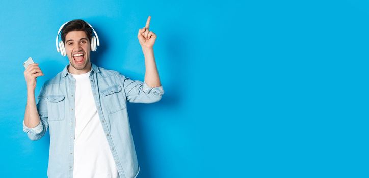 Joyful handsome man dancing with smartphone, listening music in headphones and pointing finger up, standing over blue background.