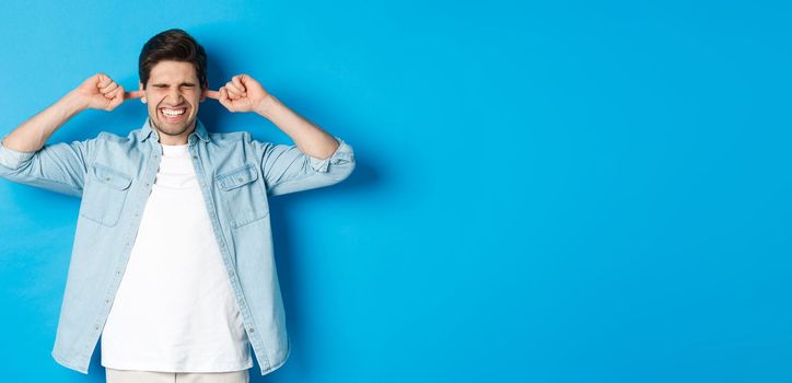 Irritated man close ears from noisy neighbours, looking bothered while standing against blue background.