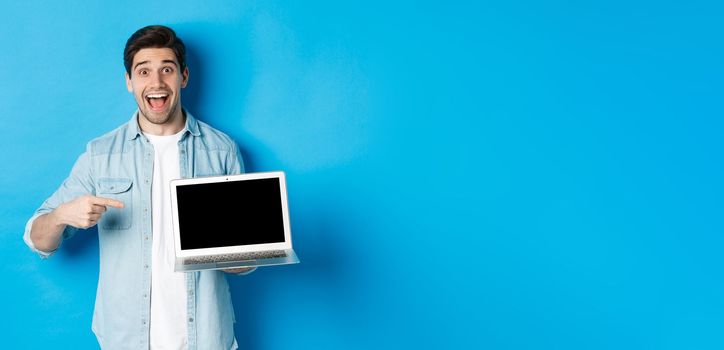 Excited smiling man pointing at laptop screen, showing internet promo offer or website advertisement, standing over blue background.