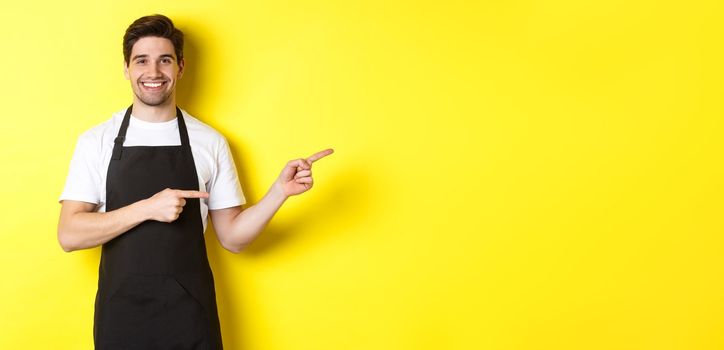 Friendly waiter pointing fingers right, showing your logo or promo offer, wearing black apron uniform, standing over yellow background,