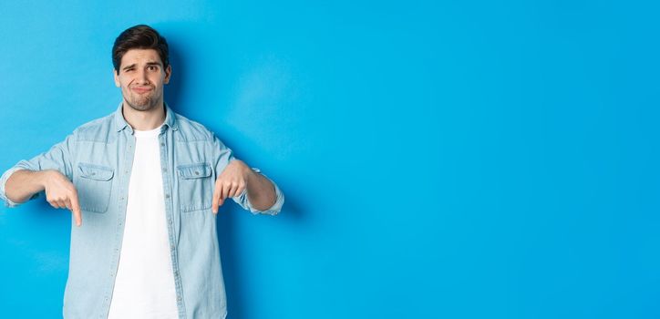 Disappointed guy in casual outfit, pointing fingers down with doubtful grimace, standing against blue background.