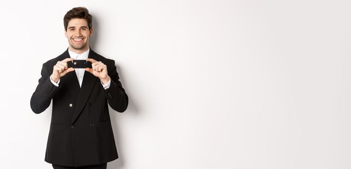 Portrait of confident handsome businessman, smiling and showing credit card, standing in suit against white background.