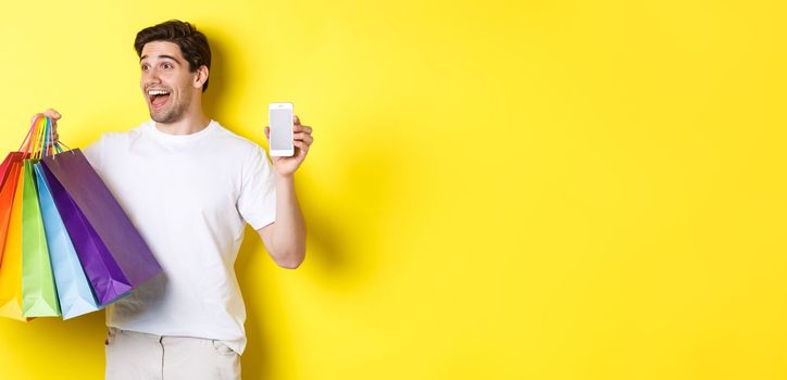 Excited man showing smartphone screen and shopping bags, achieve app goal, demonstrating mobile banking application, yellow background.