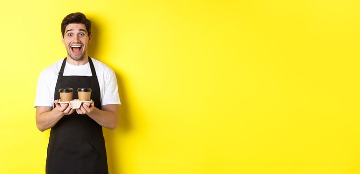 Excited male barista holding two cups of takeaway coffee, working in cafe, standing over yellow background.