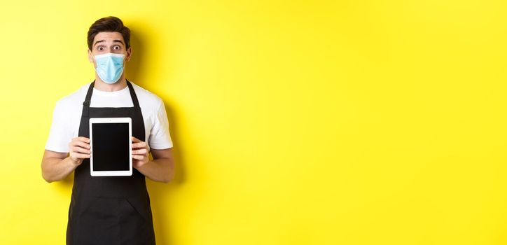 Concept of covid-19, small business and pandemic. Surprised waiter in black apron and medical mask showing tablet screen, standing over yellow background.