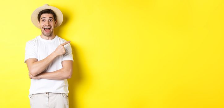 Happy male tourist in straw hat pointing finger right, showing promo offer on copy space, yellow background. Copy space