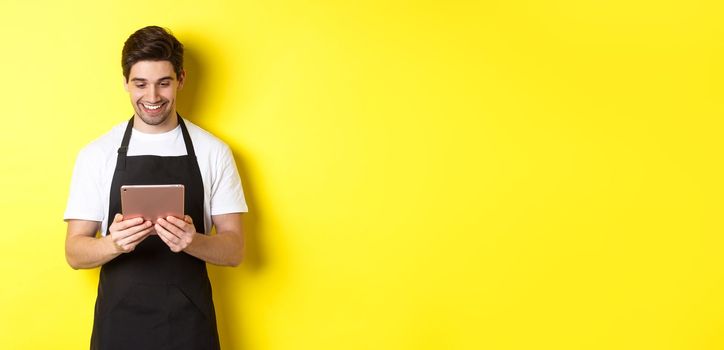 Seller in black apron looking at digital tablet screen, smiling pleased, standing over yellow background.