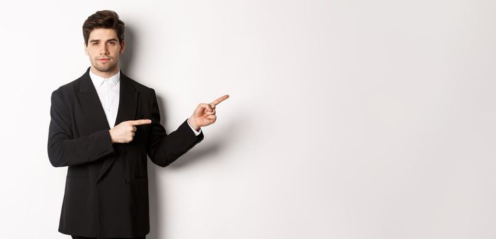 Image of handsome businessman in black suit, pointing fingers right and looking at camera, standing against white background.