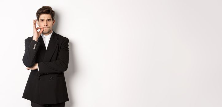 Image of serious businessman in black suit, zipping mouth and frowning, hiding secret, showing taboo sign, standing over white background.