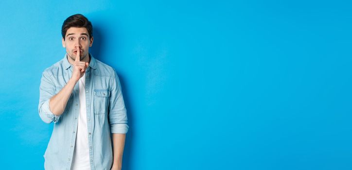 Portrait of excited man asking to keep quiet, showing hush taboo sign and looking nervously at camera, standing against blue background.