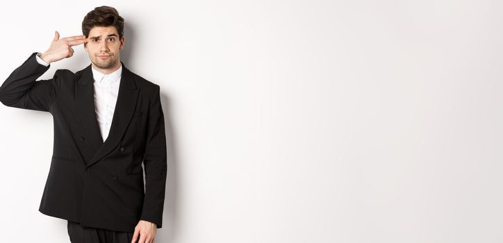 Portrait of distressed and troubled businessman in suit, pointing finger gun at head and looking bothered, shooting himself, standing against white background.