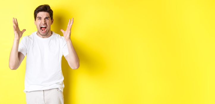 Angry man yelling and shaking hands, grimacing with hatred, standing hateful against yellow background. Copy space