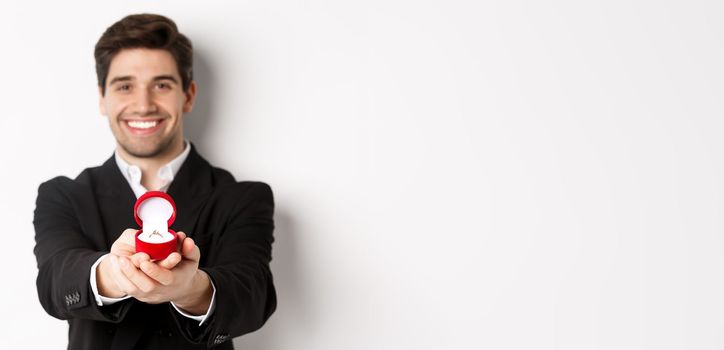 Image of handsome man looking romantic, giving you an engagement ring, making proposal to marry him, standing against white background.