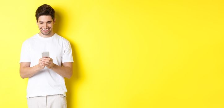 Young man reading text message on smartphone, looking at mobile phone screen and smiling, standing in white t-shirt against yellow background.