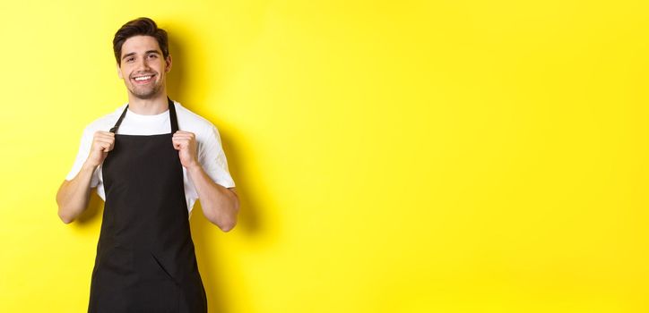 Confident barista in black apron standing against yellow background. Waiter smiling and looking happy.